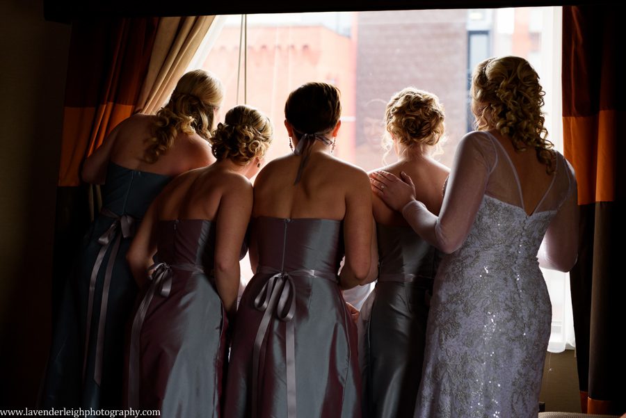 A bride and her bridesmaids peeking out the window at the groom in the street below
