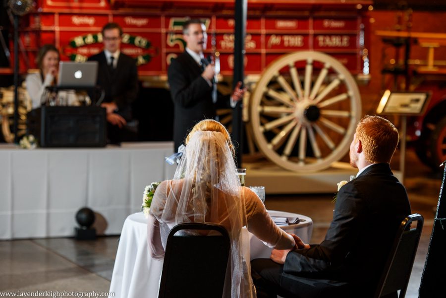 A bride and groom hold hands during the father of the bride's toast at the Heinz History Center